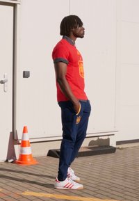 Rood T-shirt met korte mouwen en het embleem van Spanje, donkerblauwe broek met zakken en witte Adidas sneakers met rode strepen. Achtergrond is effen.
