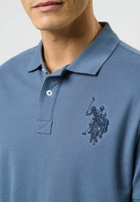 Zils polo krekls, īsas piedurknes, teksturēta auduma, ar divu pogu plāksni un izšūtu polo spēlētāja dizainu kreisajā krūtīs.