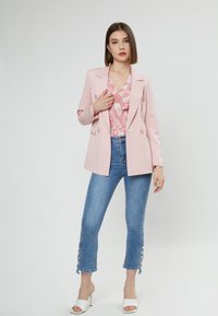 Himmelblaues Blazer mit Revers, getragen über einer floralen Bluse. Blaue Jeans in Knöchellänge mit Knopfakzenten am Saum. Weiße Absatzsandalen.