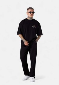 Schwarzes oversized T-Shirt mit Logo auf der Vorderseite, kombiniert mit passenden Hosen und weißen Sneakers. Glatte Stofftextur; lockere Passform. Schwarze Sonnenbrille.