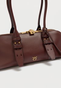 Καφέ δερμάτινη τσάντα χειρός με φερμουάρ, χρυσά μεταλλικά στοιχεία, δύο χερούλια και ρυθμιζόμενους ιμάντες. Διαθέτει ραφές και λεπτομέρεια με λογότυπο.