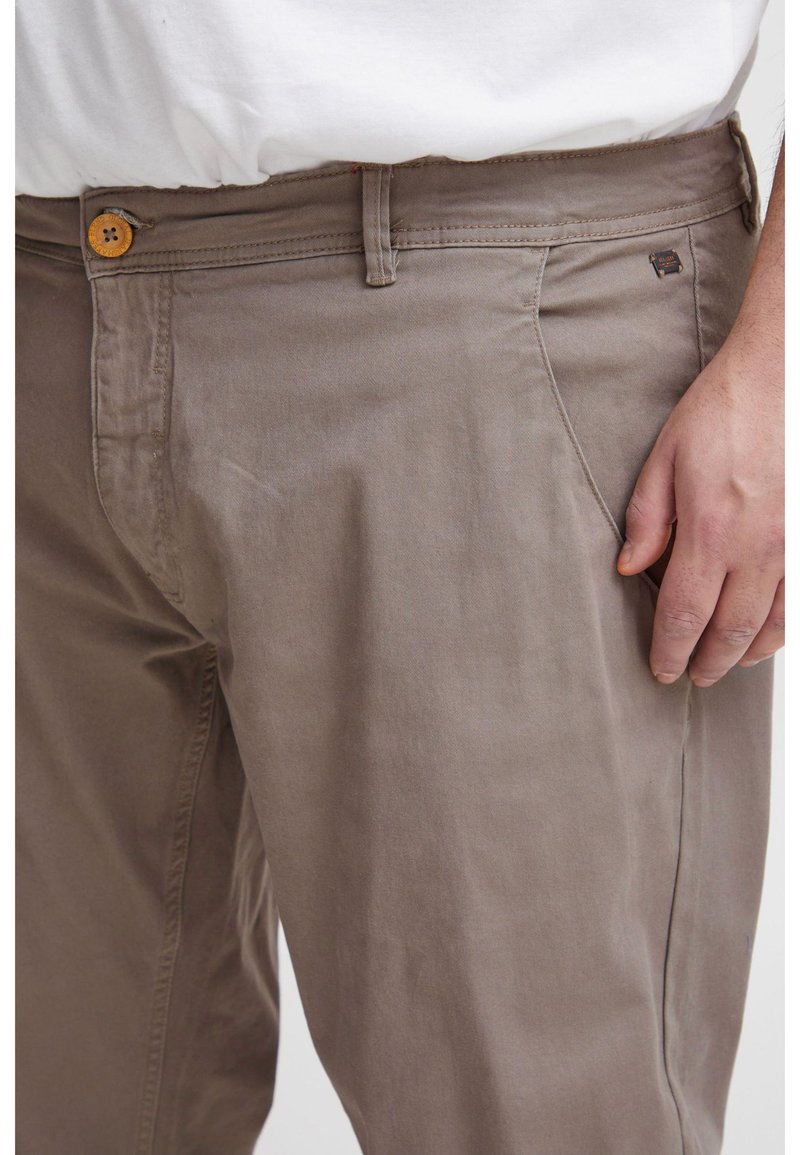 Close-up van bruine broek met knoop en riemlussen, gedragen door een persoon met de rechterhand naast de zak.