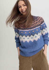 Pull en tricot présentant des nuances de bleu avec un motif fair isle en rose, blanc et orange. Manches et ourlet côtelés, porté avec un pantalon en velours côtelé.