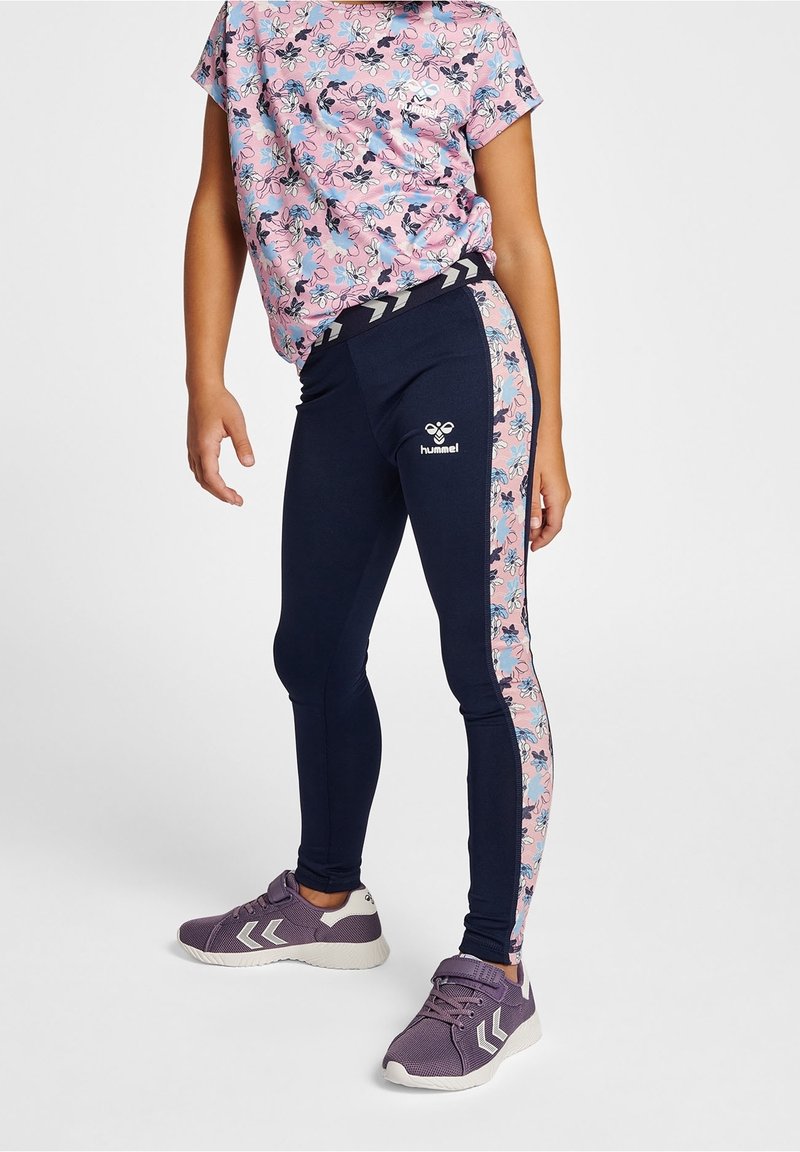 Enfant portant des leggings bleu marine avec des bandes latérales florales roses, une chemise fleurie rose et des baskets violettes, debout sur un fond uni.