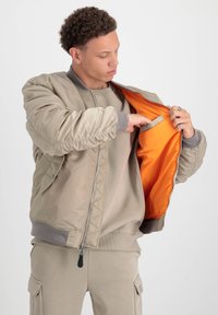 Chaqueta bomber beige con cuello acanalado y puños elásticos, con un forro naranja y un bolsillo interior. Llevada sobre un sudadera beige.
