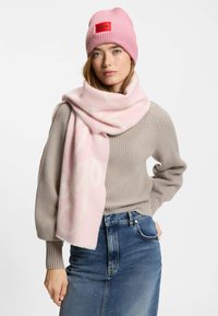 Berretto lavorato a maglia rosa con patch logo rosso; sciarpa morbida rosa chiaro con branding sottile; maglione grigio testurizzato; jeans in denim blu a vita alta.