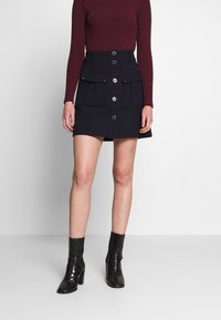 Falda azul marino de corte recto, con dos bolsillos delanteros, cierre de botones y tejido texturizado. Combinada con un top ajustado de manga larga.