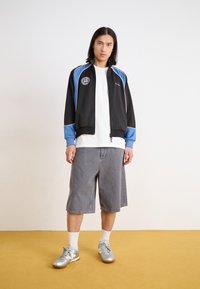 Young man standing on a yellow floor wearing a black and blue zip-up jacket, white T-shirt, gray knee-length shorts, white socks, and silver sneakers.