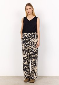 Svart ärmlös topp med V-ringning, i kombination med vida beige byxor med ett svart blommönster. Modellen står med händerna i fickorna.