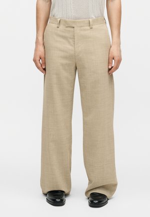 Beige bukser med vide ben, båret med sorte sko, sat sammen med en hvid strikket top med korte ærmer, stående mod en ensfarvet baggrund.