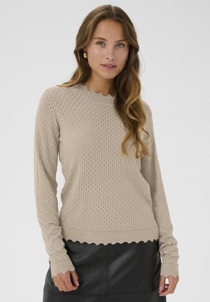Beige gebreide trui met een textuurpatroon, golvende halslijn en omgeslagen mouwen, gecombineerd met een zwarte leren rok.