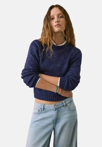 Granatowy sweter dzianinowy z fakturowanym wzorem i prążkowanym dołem, nałożony na białą koszulę z kołnierzykiem, zestawiony z jasnoniebieskimi spodniami typu wide-leg.