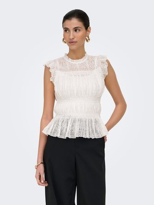 Donna che indossa un top peplum bianco in pizzo senza maniche con collo alto arricciato e pantaloni neri, in piedi con la mano in tasca.