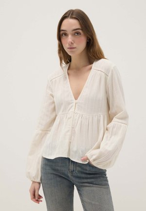 Jeune femme aux longs cheveux bruns portant un chemisier ample de couleur crème avec boutons et manches bouffantes, associé à un jean bleu, en position neutre.