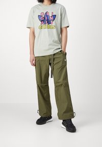 Světle zelené tričko s grafickým designem "ORIGINALS adidas"; olivově zelené cargo kalhoty s nastavitelným pasem; černé sportovní boty.