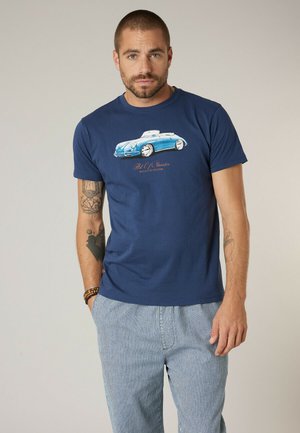 Homme portant un T-shirt bleu marine avec un motif de voiture classique bleue et un pantalon rayé bleu clair et blanc, debout devant un fond uni.