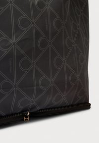 Čierna textilná taška s geometrickým vzorom prepletených slučiek a značkovým logom. Obsahuje viditeľný zips pozdĺž spodnej časti.