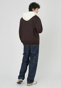 Veste marron avec capuche blanche, assortie à un pantalon cargo bleu foncé ample. Caractéristiques : poignets élastiques et plusieurs poches.