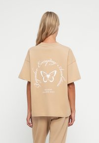 Béžové oversized tričko vyrobené z bavlny, na zadn�í straně s grafikou bílé motýlice a textem. Krátké rukávy a kulatý výstřih.