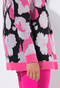 Schwarzer Strickpullover mit pink-weißen Blumenmustern und einem gerippten rosa Saum. Das Model trägt rosa Hosen, mit einer sichtbaren Hand.