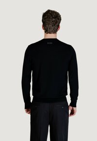 Maglione nero con scollo tondo, polsini e orlo a coste, con un piccolo logo sulla parte superiore della schiena. Indossato con pantaloni neri.