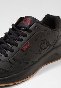 Schwarze Sportsneaker mit Obermaterial aus synthetischem Leder, schwarzen Schnürsenkeln, roten Logo-Akzenten und einer Gummisohle mit strukturiertem Muster.