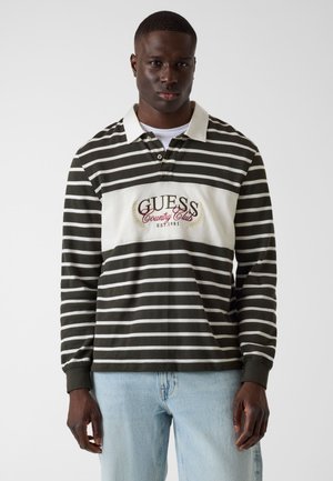 Homme portant une chemise à manches longues rayée vert foncé et blanc avec le logo "Guess Country Club", et un jean bleu clair, debout devant un fond uni.