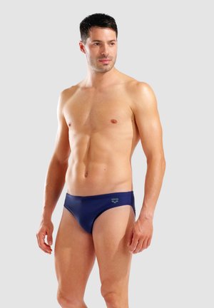 Uomo in forma in piedi, indossa slip da bagno blu navy, guarda leggermente di lato su uno sfondo grigio chiaro uniforme.