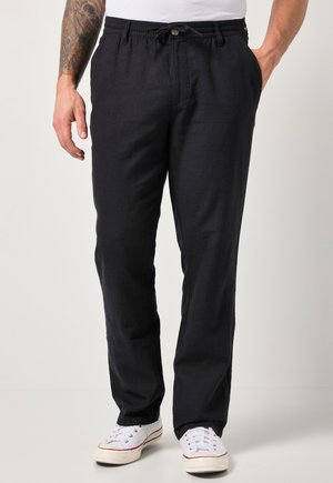 Hombre con pantalones negros con cordón y zapatillas blancas, de pie con las manos en los bolsillos, tatuaje visible en el antebrazo izquierdo.