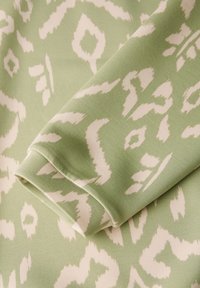 Vihreä kangas, jossa on pehmeä tekstuuri ja valkoinen eläinkuosikuvio. Kangas on rullattu yhdestä reunasta, mikä paljastaa sen sileän pinnan.