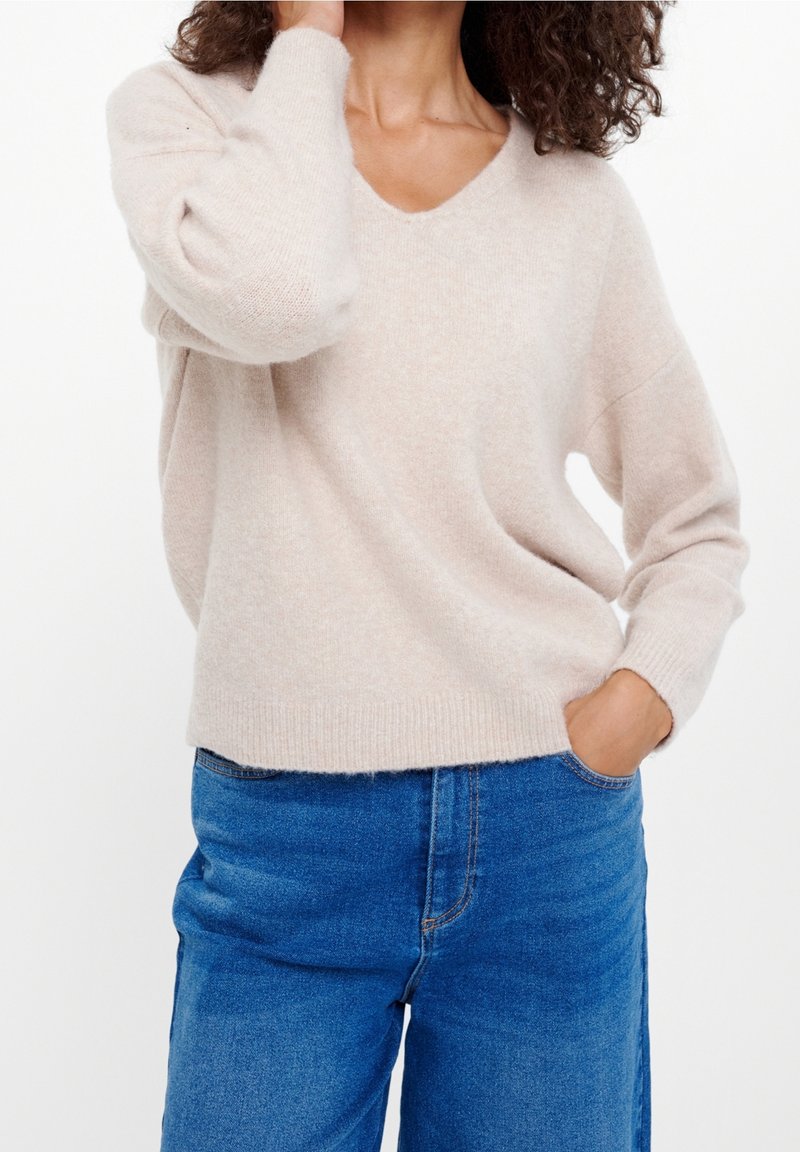 Femme portant un pull à manches longues beige clair et un jean bleu, une main dans la poche et l’autre touchant ses cheveux.