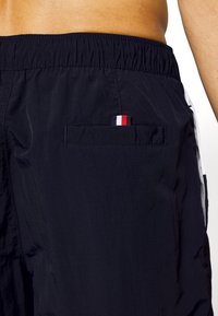 Shorts da bagno blu navy con vita elasticizzata; presenta una tasca posteriore con un piccolo etichetta rossa, bianca e blu. Tessuto liscio e leggero.