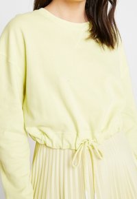 Top giallo a maniche lunghe e a coste con vita regolabile. Presenta una vestibilità rilassata ed è abbinato a una gonna plissettata di un giallo chiaro.