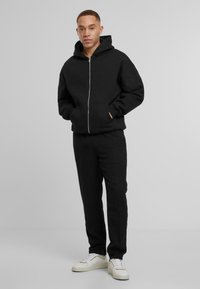 Schwarzer Reißverschluss-Hoodie mit Kängurutasche, kombiniert mit passenden Hosen. Das Outfit hat eine entspannte Passform und zeigt eine glatte Textur. Akzentuiert mit weißen Sneakers.