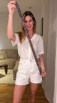 Hochgehaltener Gürtel aus Stoff im Leopardenmuster, getragen zu einem weißen Hemd mit Kragen und weißen Shorts. Im Hintergrund sind ein beiges Sofa und ein Holzboden zu sehen.