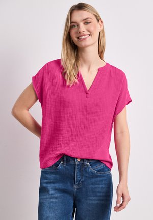 Uśmiechnięta kobieta z długimi blond włosami nosi jasnoróżową teksturowaną bluzkę z krótkim rękawem i niebieskie jeansy, stojąc na jednolitym tle.