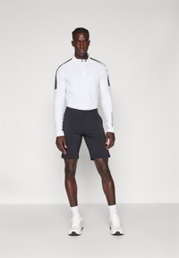 Hvit langermet zip-up skjorte med svarte detaljer, kombinert med svarte shorts. Modellen har på seg hvite joggesko og hvite sokker. Enkelt design.