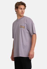 Purple cotton t-shirt with short sleeves, oversized fit, featuring a colorful graphic logo on the left chest. Smooth texture, basic round neckline.