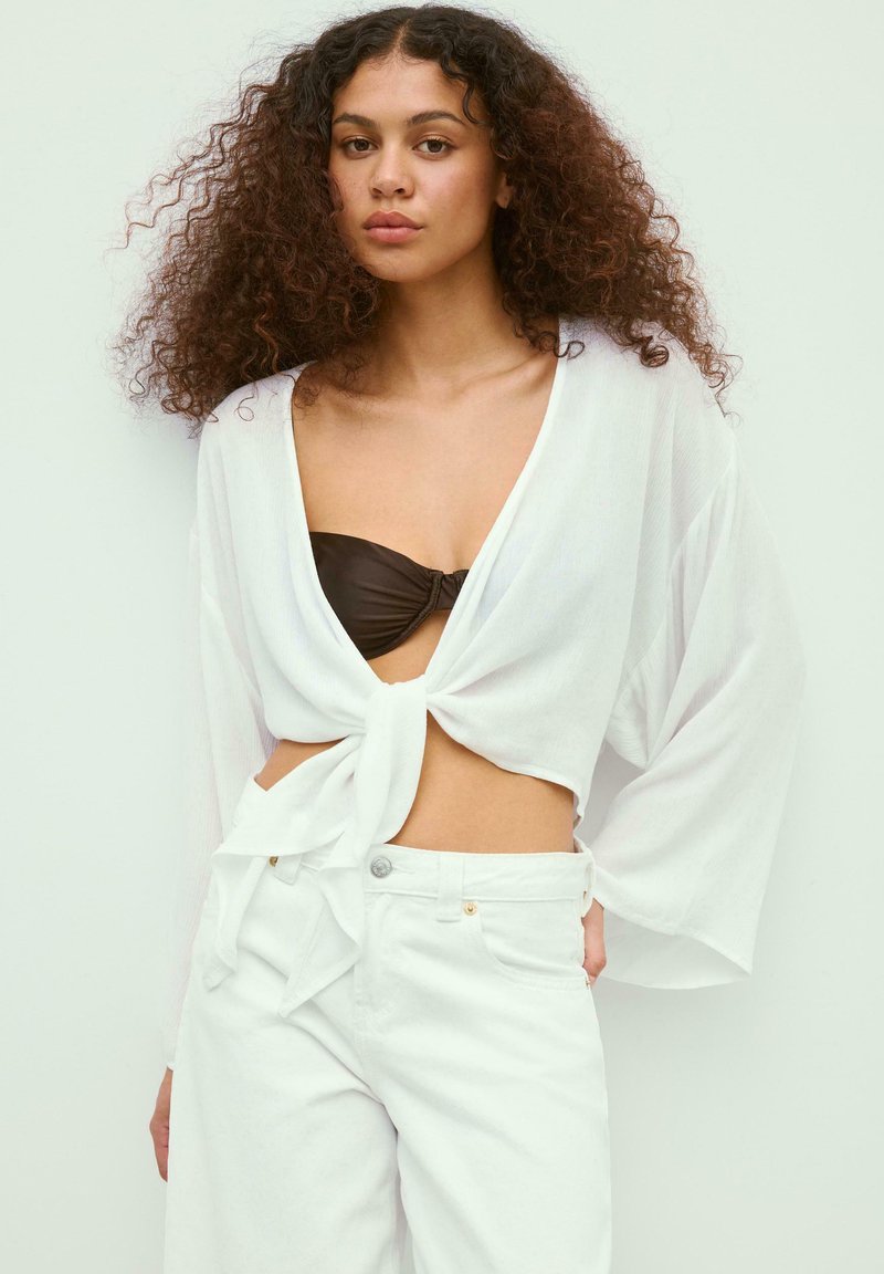 Jeune femme aux cheveux bouclés porte un chemisier blanc noué devant, par-dessus un bandeau noir et un pantalon blanc taille haute, debout devant un fond uni.