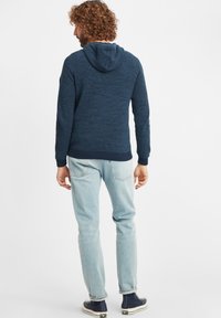 Blaue Strickkapuzenjacke mit strukturiertem Finish, ausgestattet mit einer Kapuze sowie gerippten Bündchen und Saum, kombiniert mit hellen Jeans und dunklen Sneakers.