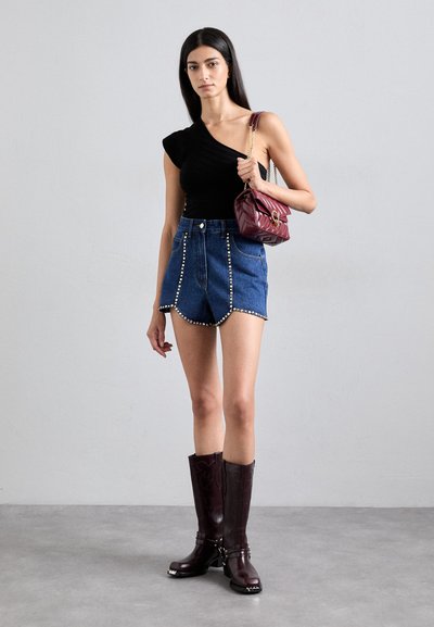Top nero a spalla unica, pantaloni shorts in denim a vita alta con dettagli in borchie metalliche, borsa a tracolla bordeaux scuro e stivali da cowboy marroni lucidi.