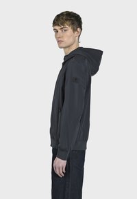 Chaqueta ligera de color gris oscuro con capucha, bolsillos frontales con cremallera, puños elásticos y un acabado de tela texturizada. Bajo ajustable para un mejor ajuste.