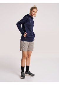 Marineblauer Hoodie mit Frontlogo, graue Shorts mit schwarzen Akzenten und seitlichen Reißverschlüssen, schwarze Knöchelsocken und graue Sportschuhe.