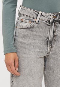 Jeans de mezclilla gris de cintura alta con un acabado desgastado. Presentan bolsillos frontales y herrajes de cobre, con detalles de costura visibles.
