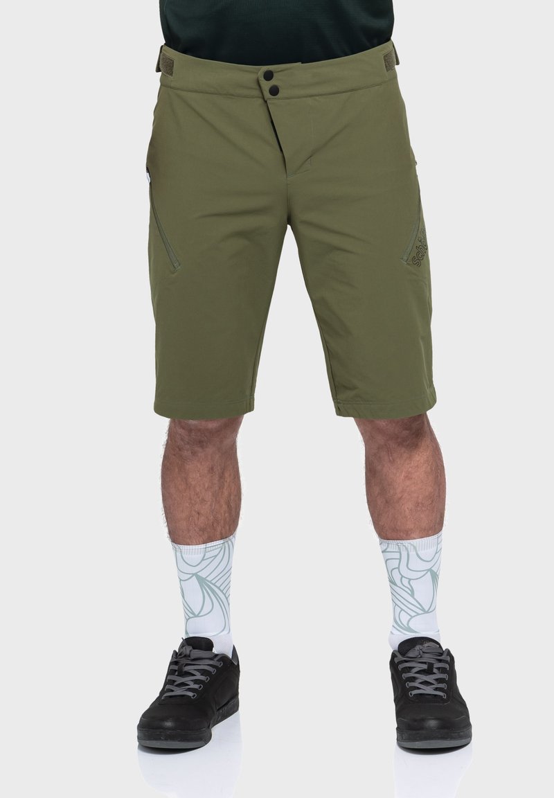 Olivgrüne Shorts mit flachem Schnitt, zwei mit Reißverschluss versehenen Seitentaschen und Logostick. Kombiniert mit schwarzen Sneakers und gemusterten weißen Socken.
