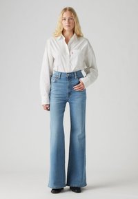 Fehér hosszú ujjú, gombos ing zsebbel, világoskék, magasderekú trapéznadrággal párosítva. A modell fekete cipőt visel. Egyszerű háttér.