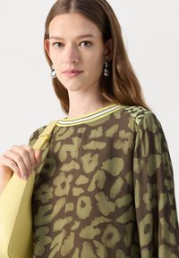 Bruine blouse met groen abstract luipaardpatroon, ronde halslijn met gele strepen, gecombineerd met een gele leren tas. Metallic oorbellen zichtbaar.