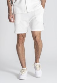 Pantalones cortos blancos texturizados con un corte corto, combinados con zapatillas blancas con cordones gruesos. Diseño minimalista y líneas limpias. Etiqueta negra en los pantalones cortos.