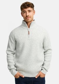 Mand med lyst brunt hår iført en lysegrå ribbet zip-up sweater og mørkeblå jeans, hænderne i lommerne, neutral baggrund.
