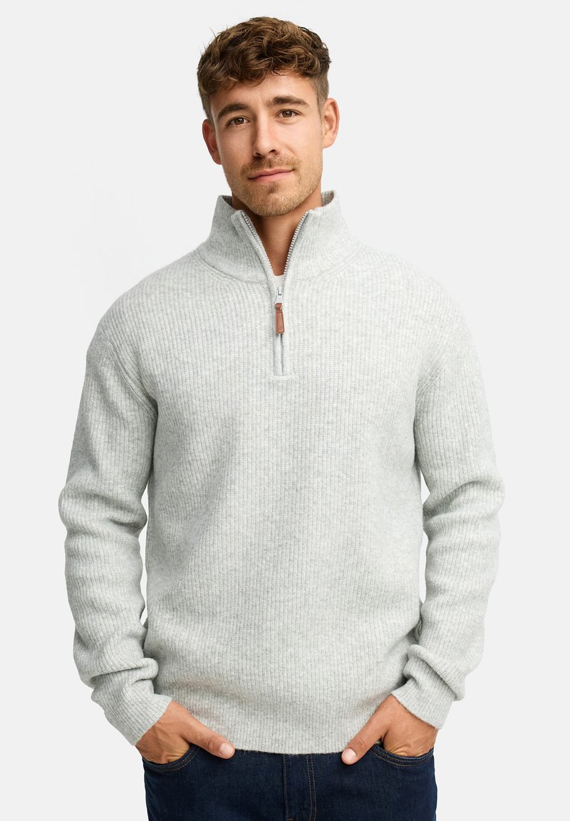 Mand med lyst brunt hår iført en lysegrå ribbet zip-up sweater og mørkeblå jeans, hænderne i lommerne, neutral baggrund.
